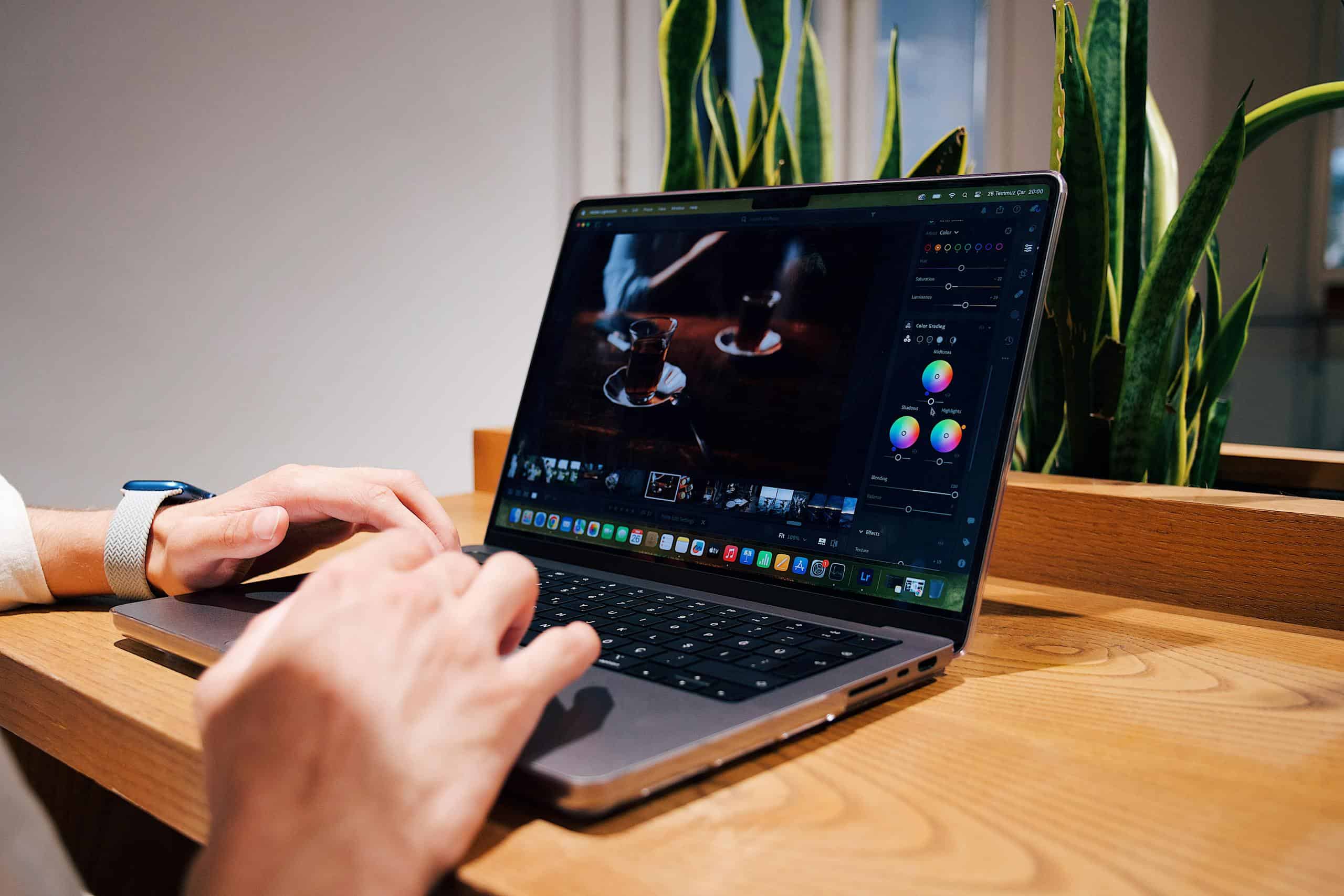 Close-up of hands editing a photo on a laptop in an office setting, surrounded by green plants.